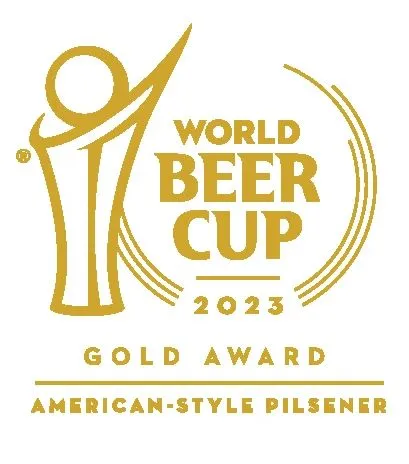 The 2023 World Beer Cup® Gold Medal awarded to von C Brewing Co. for O.G. Pils in the American-Style Pilsener category — embossed brass medallion with the Brewers Association seal, mounted on the white-brick wall of the Norristown taproom