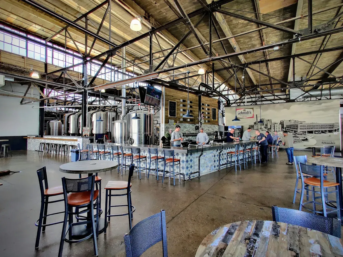 Wide interior view of the von C Brewing Co. taproom in Norristown — full bar at center with patterned blue-and-white tile front, stainless brewhouse tanks visible to the left, exposed steel-truss warehouse ceiling, Schmidt's of Philadelphia archival mural anchoring the right wall, customers seated at the bar
