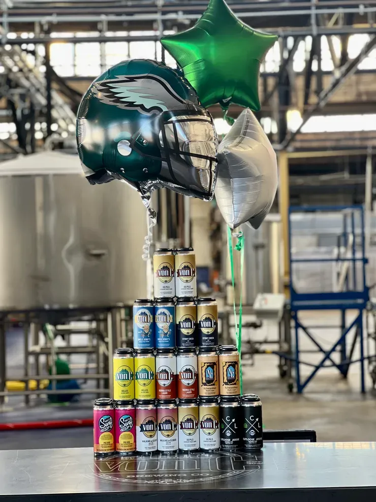 Stacked pyramid of every von C Brewing Co. 16-oz can SKU on the brewery floor — O.G. Pils, Uncle Jerry's Wit, Stony Creek IPA, Helles, Vienna Style Lager, Schwarzbier, Walking on Clouds, Pig Dog Pale Ale, and CutLoose Joose — with Philadelphia Eagles helmet and star foil balloons floating above against the open production-floor backdrop