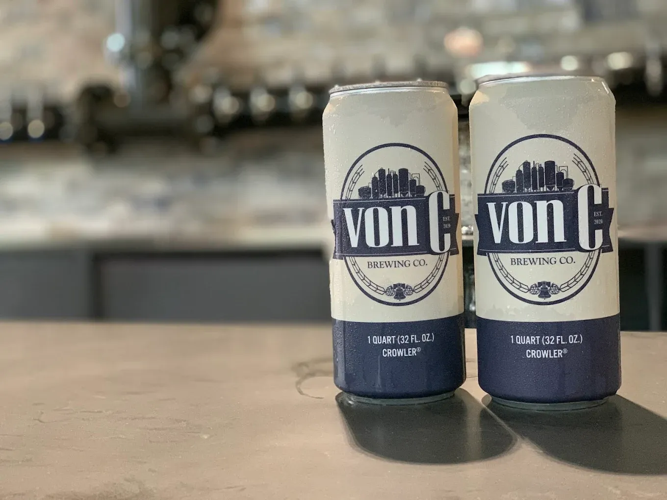 Two 32-oz crowlers with von C Brewing Co. branding standing on the stainless bar — fresh-filled fill-to-order packaging available for taprooms and bottle shops alongside the canned lineup
