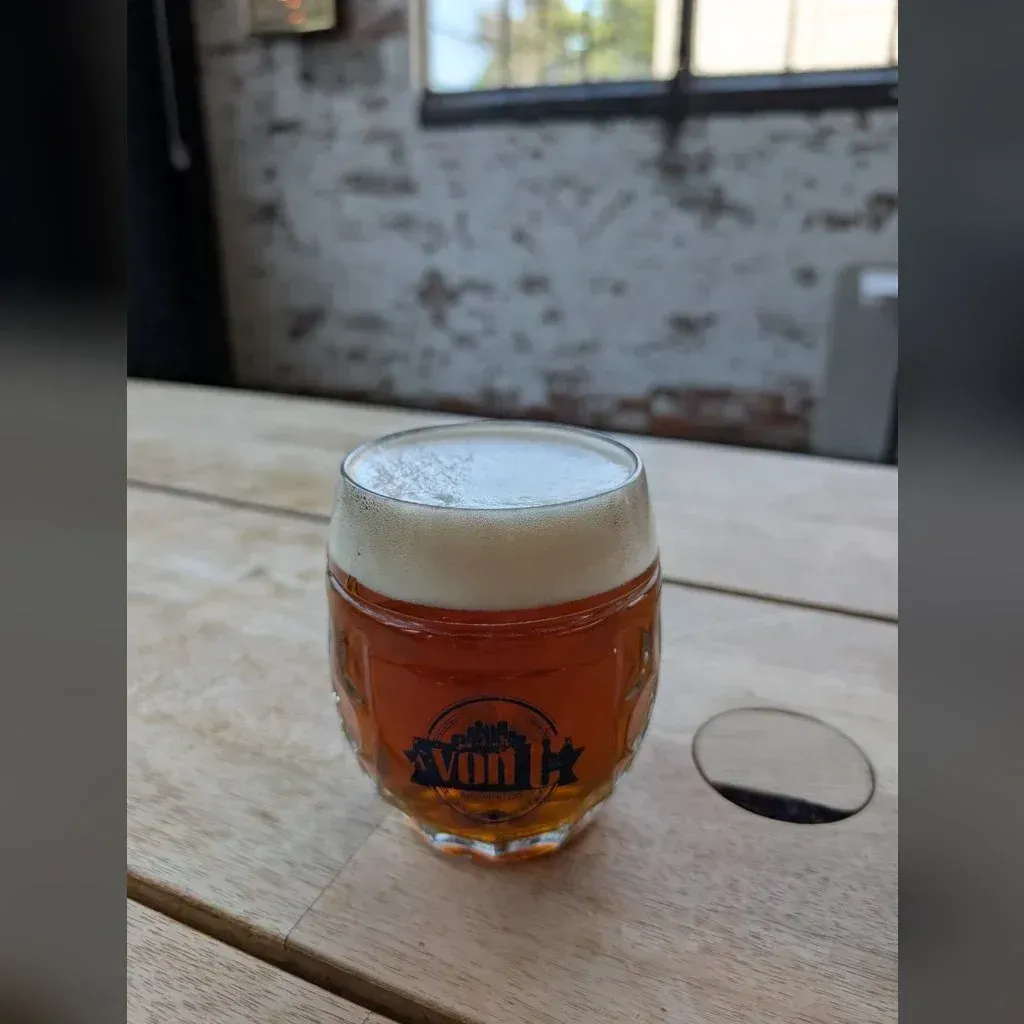 Copper-colored Czech amber lager in a von C Brewing branded dimpled mug on a wooden bar top in the Norristown taproom, cream head and brick wall behind