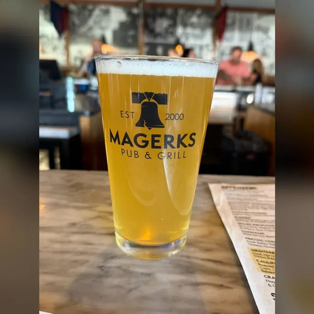 Hazy yellow Dolcita pale ale in a tall pint glass on a metal bar top — light gold with veiled clarity, soft head, indoor warm light