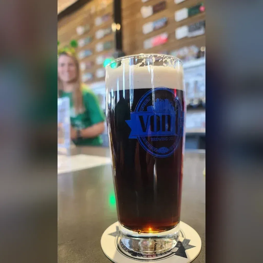 Dark amber-to-black pour of Double Haul American Stout in a von C Brewing branded pint glass on the bar at von C Brewing Co. in Norristown PA, a friend in green visible in the soft background