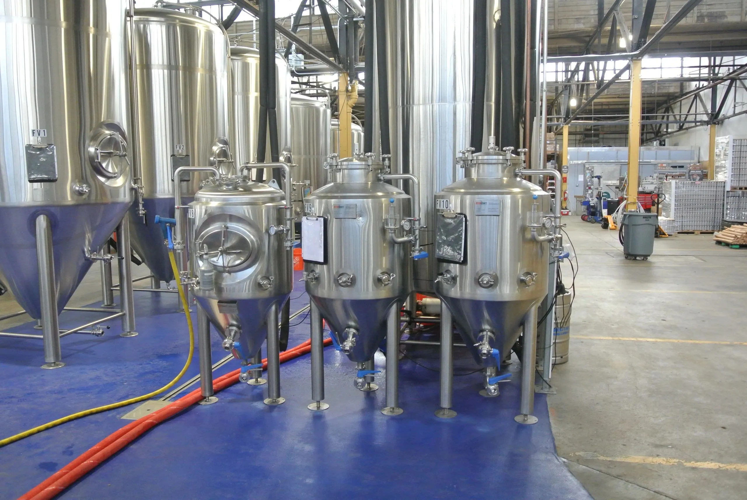 Conditioning tanks lined up on the production floor at von C Brewing Co. in Norristown PA — stainless-steel cylindroconicals in the foreground, larger fermentation tanks rising behind them, the row where every lager spends four weeks cold before service