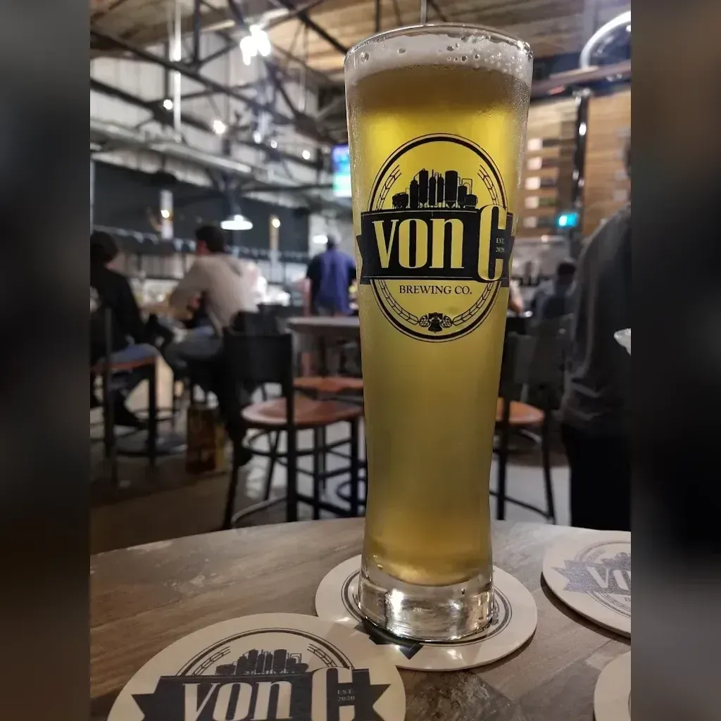 Pale gold Fräulein German pilsner in a von C Brewing branded pilsner glass on a coaster at the Norristown taproom bar, taps and bunting visible behind