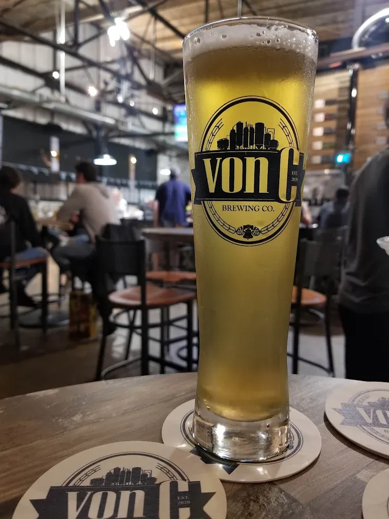 Custom vonC Brewing Co. dimpled glass mug holding a freshly poured lager at the taproom bar — textured glass faceting catching warm tungsten light, the traditional German krug shape used exclusively for the lager lineup