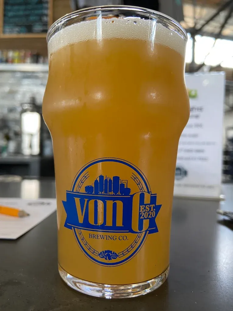 Hazy yellow-orange NEIPA in a branded von C Brewing pint glass, dense lacing around the rim, warm taproom lighting and copper bar accents behind