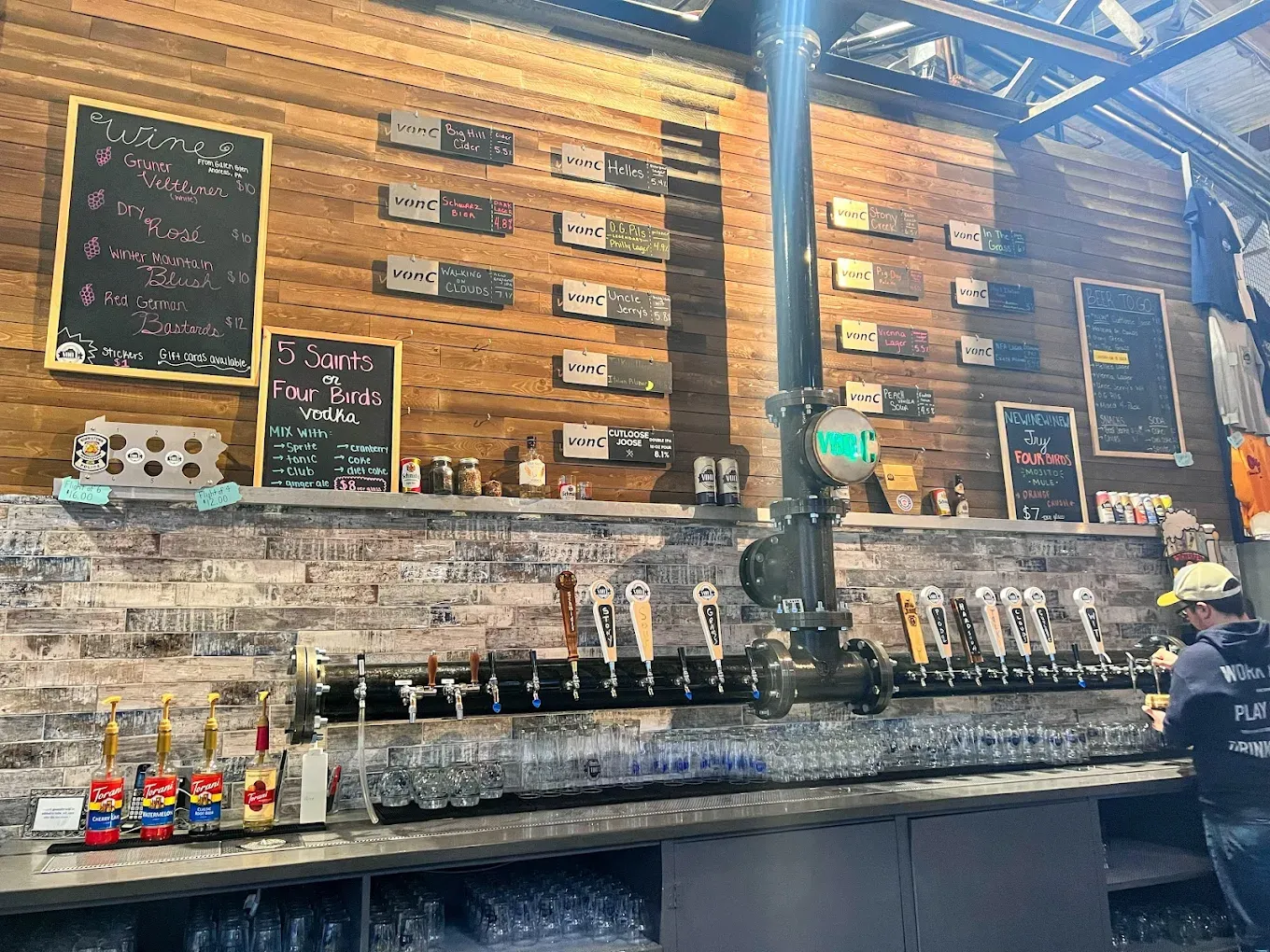 Tap wall close-up at von C Brewing with 14 brass tap handles in a row, chalkboard tap list above showing beer names and ABVs