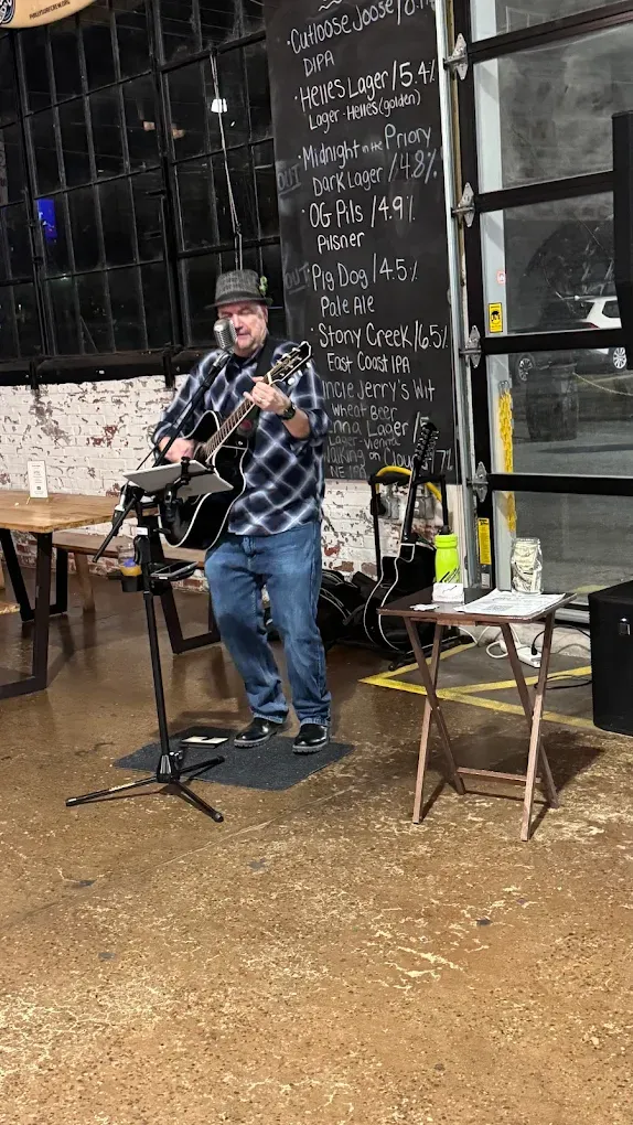 Solo acoustic performer at the corner of the von C Brewing Co. taproom in Norristown PA — guitar in hand, microphone stand at chest height, the chalkboard rotating-tap list visible on the wall behind, taproom guests turned to listen from the bar