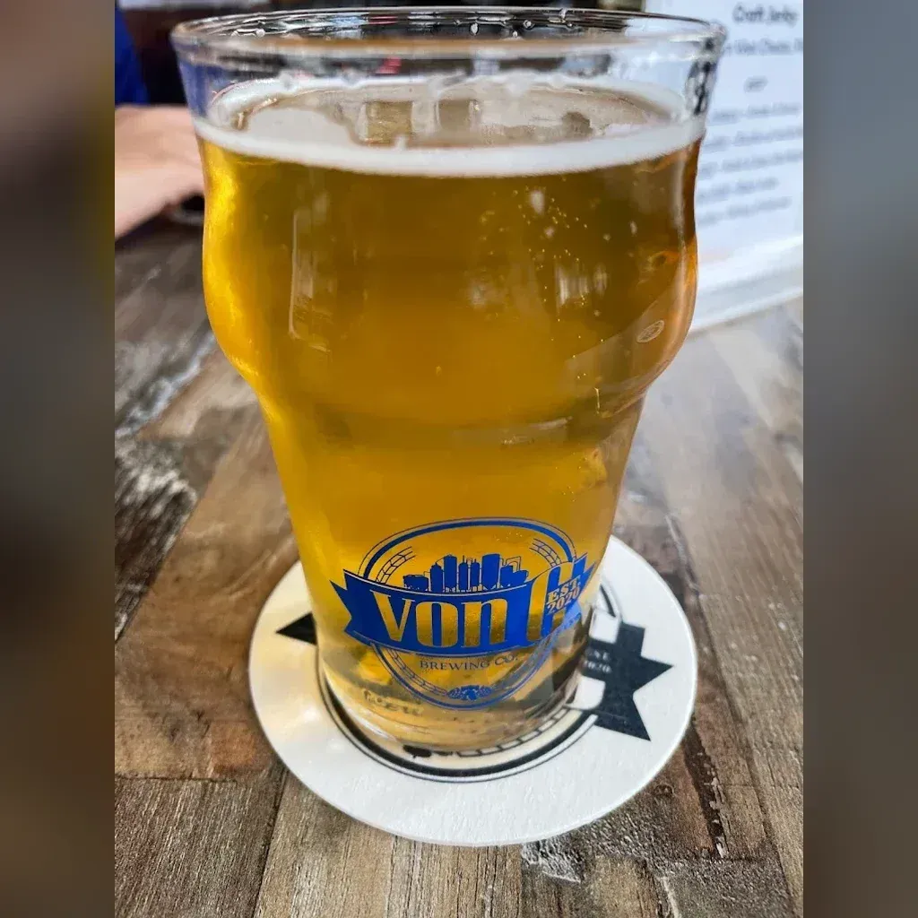 Branded von C Brewing pint glass of pale-gold Helles Lager on a reclaimed-wood table at the Norristown taproom, foamy crown, the blue vonC ribbon mark prominent on the glass