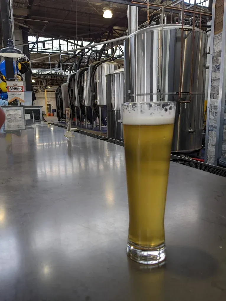 Tall slim pilsner glass of pale-gold Helles Lager on the bar at von C Brewing Co. in Norristown PA, the production-floor row of stainless fermentation tanks running into the background
