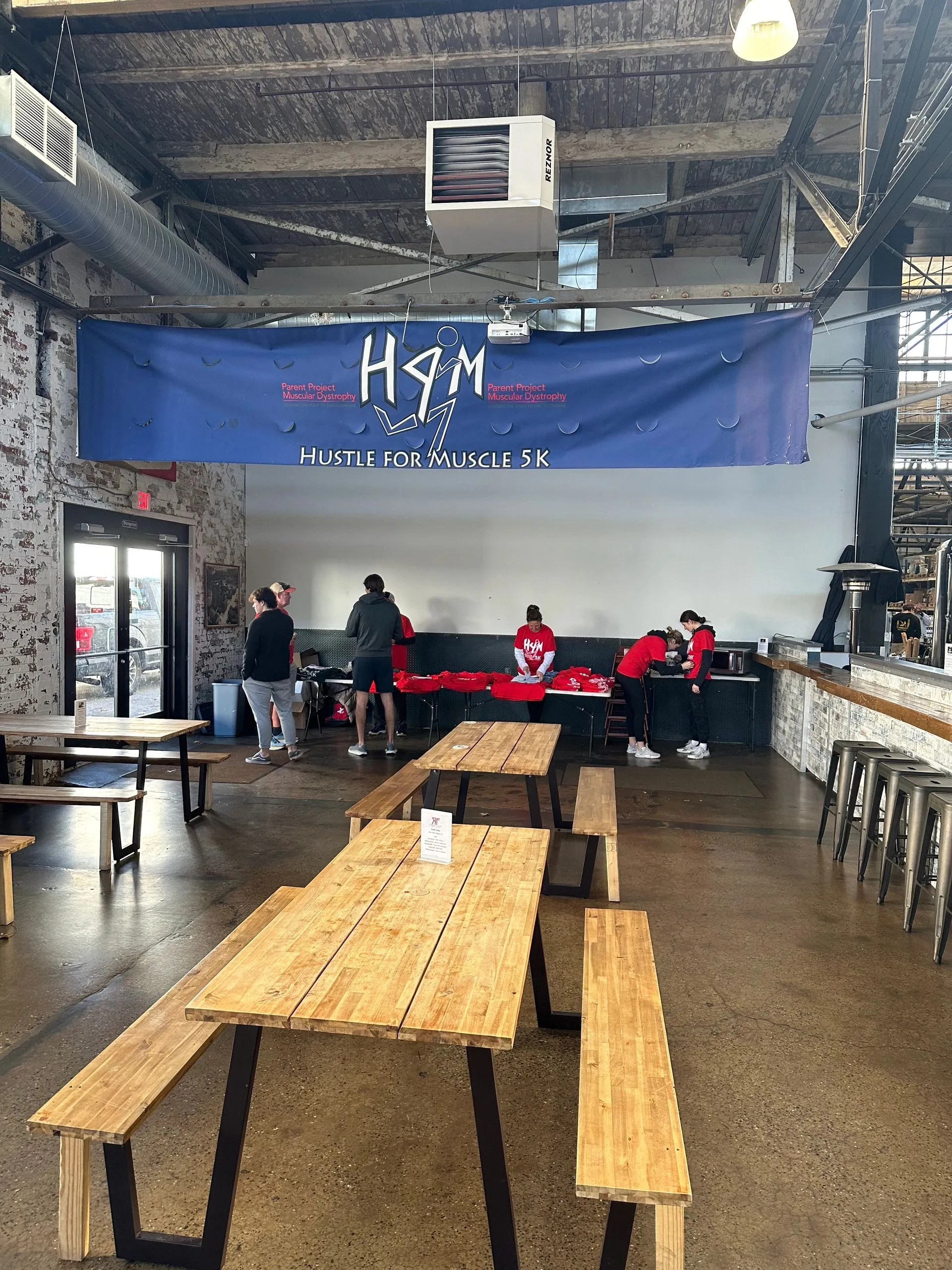 Private-event setup at the von C Brewing Co. beer garden in Norristown PA — long picnic tables arranged for a 5K-style event, signage staked at the perimeter, polished concrete floor and warehouse-roof trusses overhead, the room set for partial buyout
