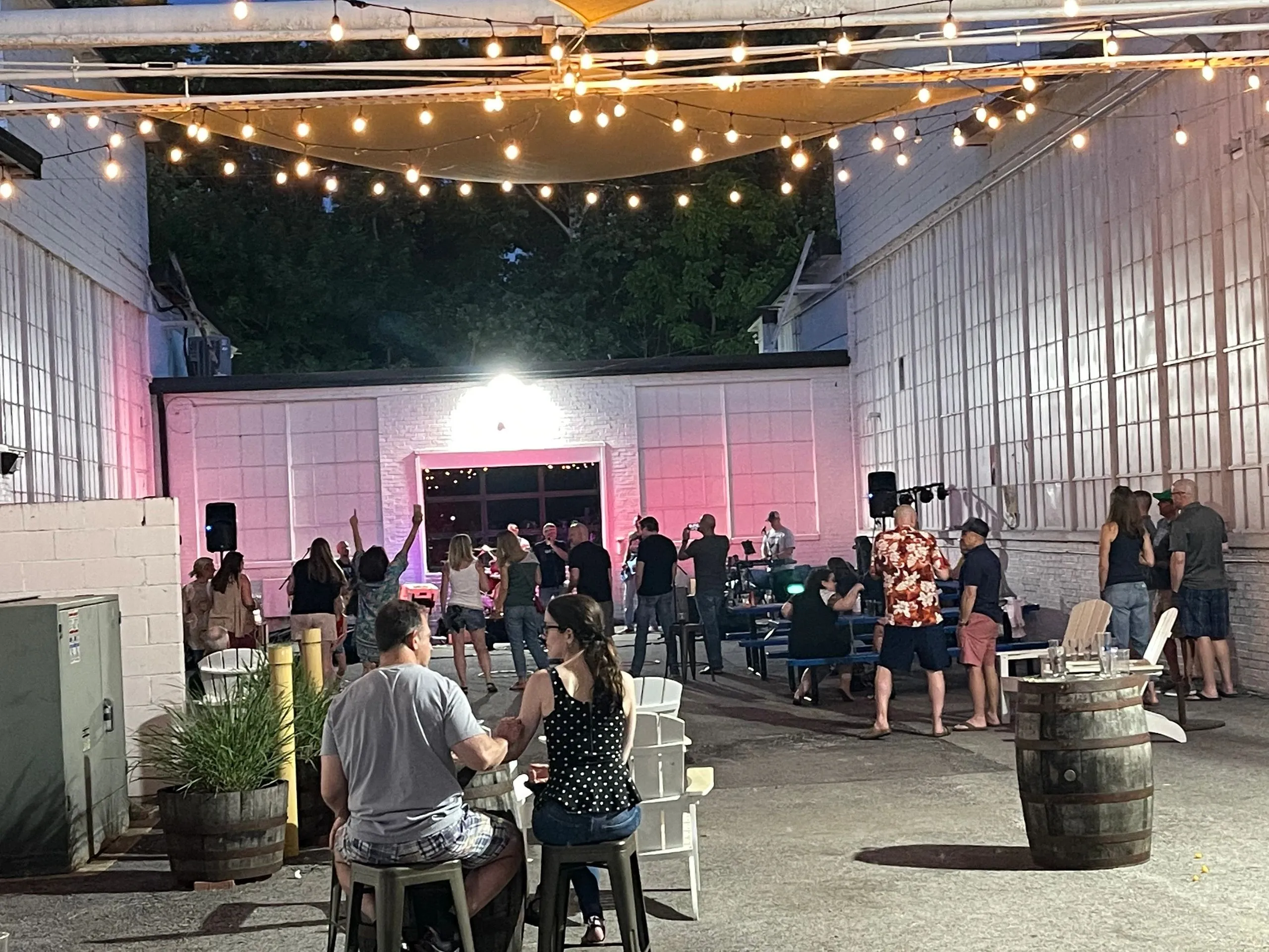 The von C Brewing Co. beer garden at night with strings of warm Edison bulbs criss-crossing overhead — picnic tables filled with seated guests, live music visible at the back of the garden, the reclaimed-warehouse facade glowing in the background