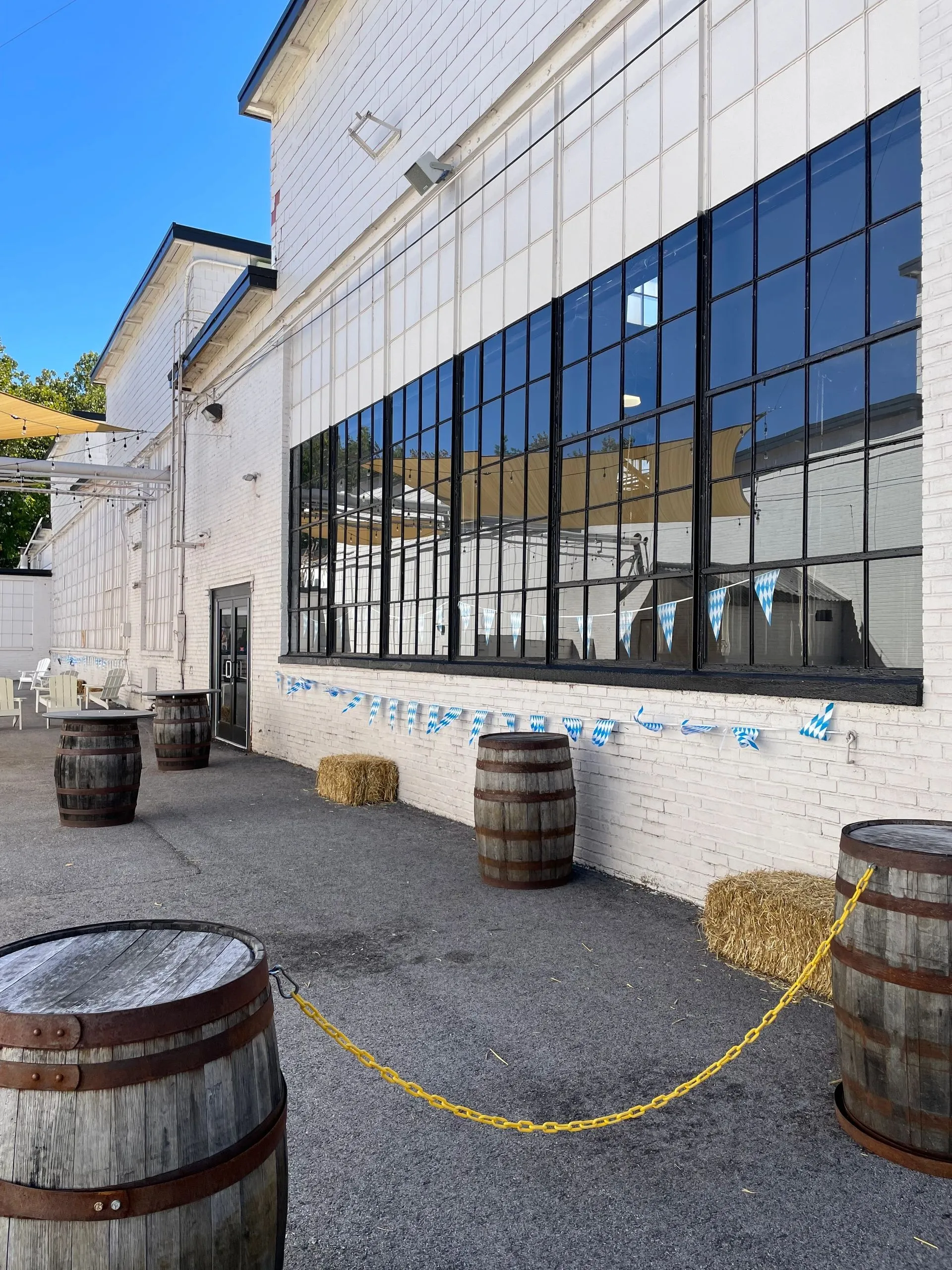 Daytime beer garden set up for Oktoberfest at von C Brewing Co. — blue-and-white Bavarian banners strung between posts, wooden barrels staged along the perimeter, families and dog-walkers seated at picnic tables in the open-air gathering