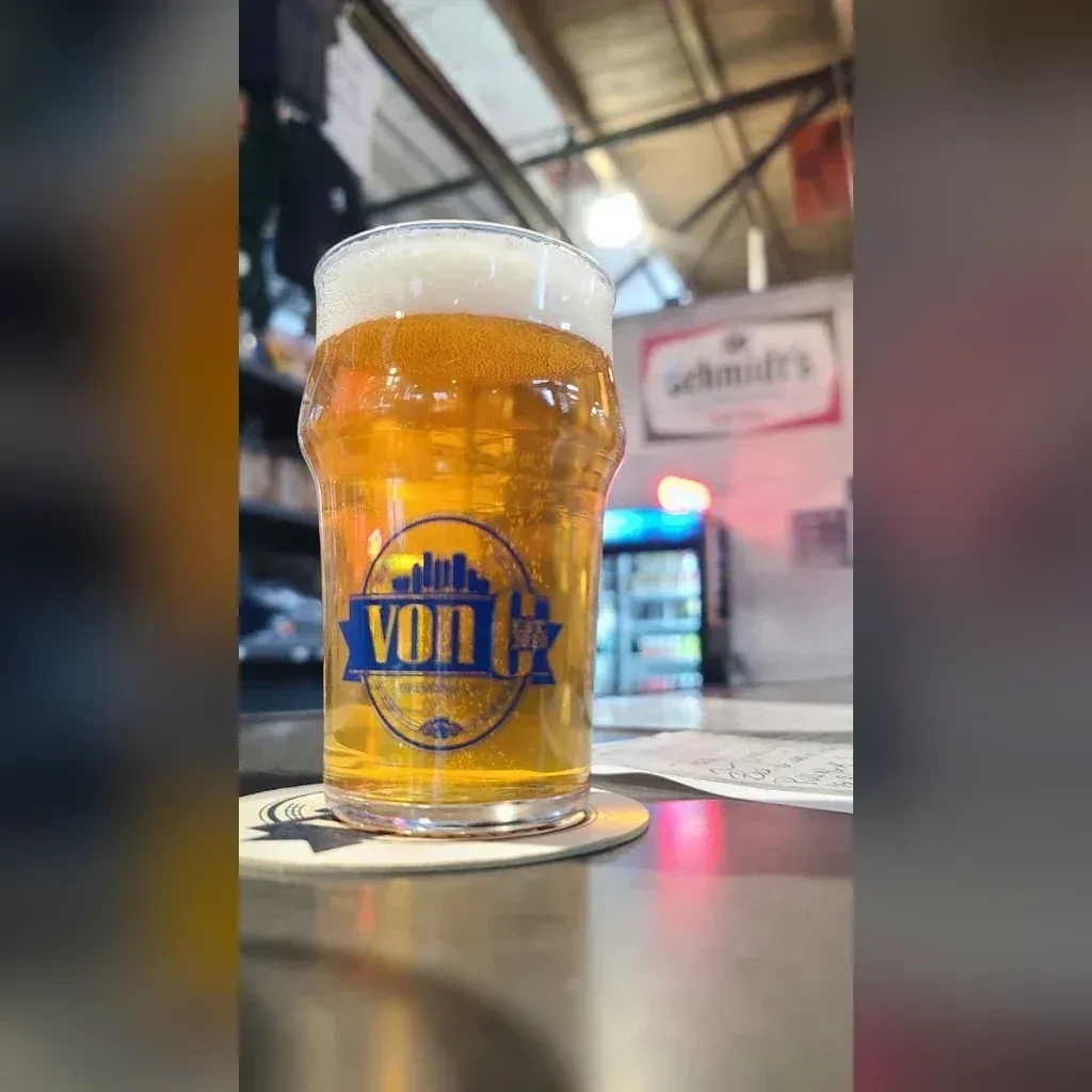 Pale amber pour of In the Grass West Coast Pale Ale in a von C Brewing branded pint glass on the bar, the Schmidt's-of-Philadelphia heritage signage visible in the background