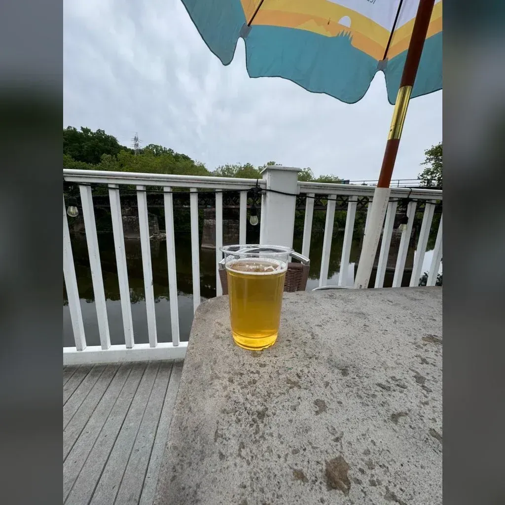 Pale gold Lime Danny light lager outdoors on a wooden deck rail at von C Brewing Norristown, river and trees visible behind under a soft umbrella, summer afternoon light