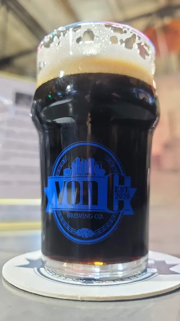 Deep mahogany Midnight in the Priory Schwarzbier in a von C Brewing branded pint glass with a dense tan-cream head, taproom interior softly behind — the German dark-lager tradition on a rotating tap