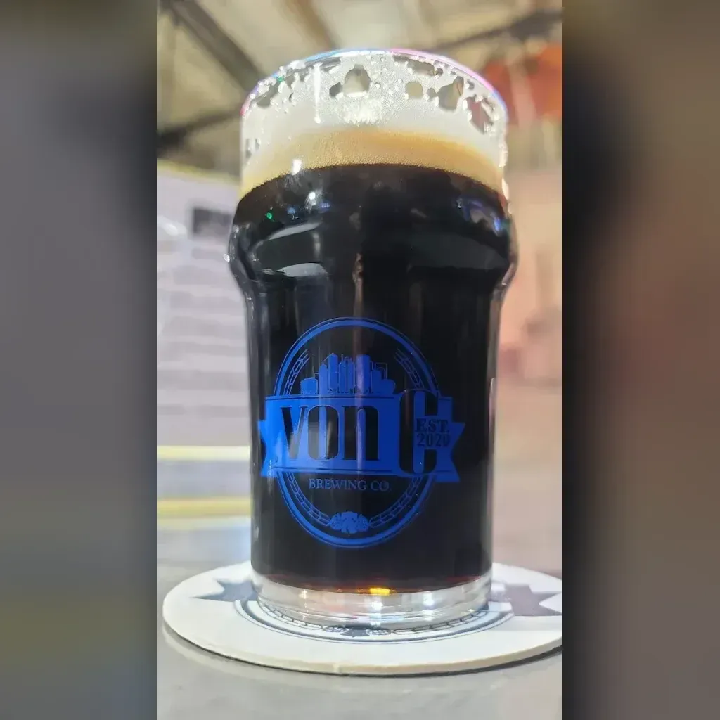 Deep mahogany Midnight in the Priory Schwarzbier in a von C Brewing branded pint glass with a dense tan-cream head, taproom interior softly behind — the German dark-lager tradition on a rotating tap