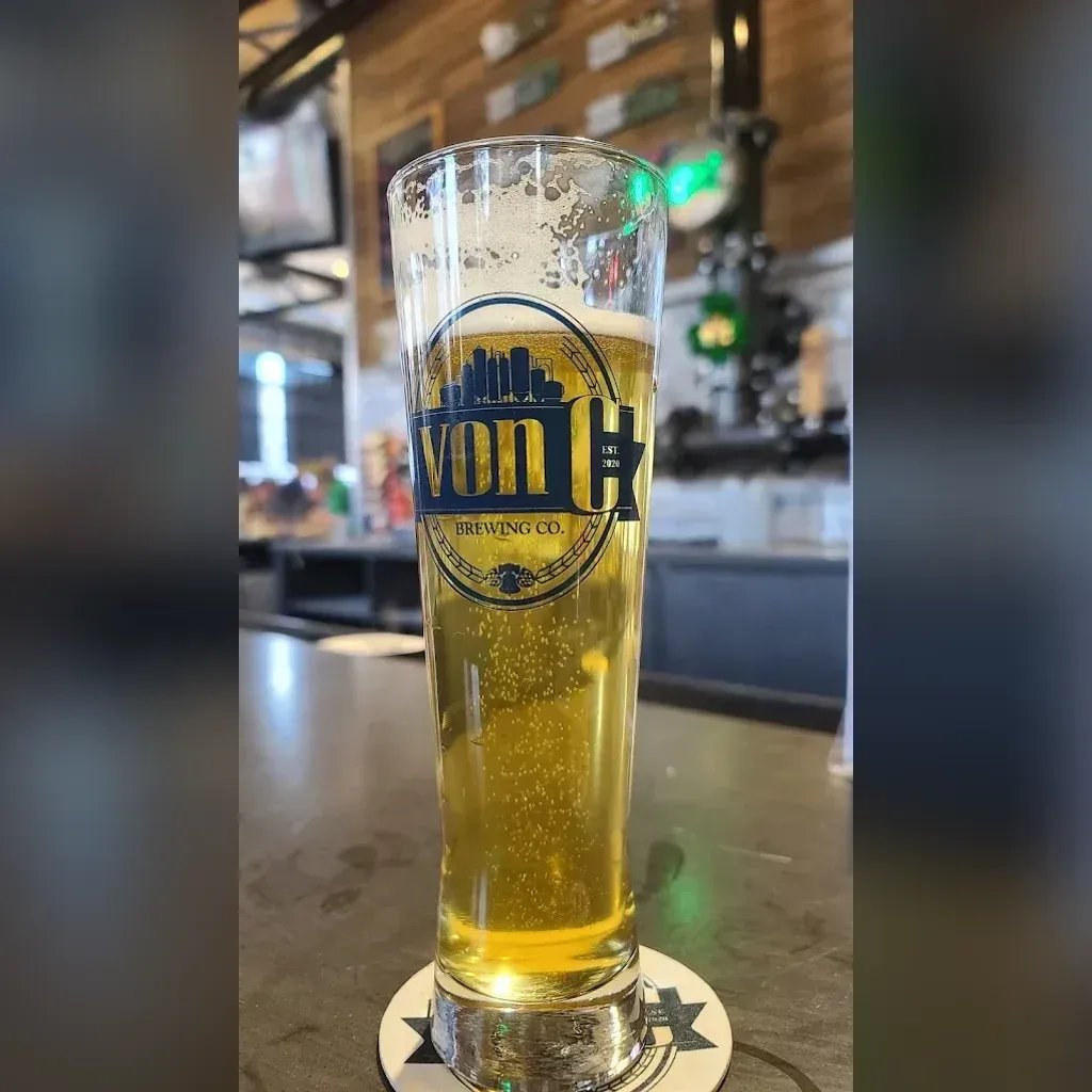 Tall pilsner pint of O.G. Pils on the von C Brewing Co. bar in Norristown, condensation beading on the glass, full fermenter line and Schmidt heritage signage visible in the background