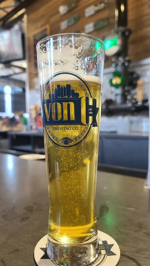 Tall pilsner pint of O.G. Pils on the von C Brewing Co. bar in Norristown, condensation beading on the glass, full fermenter line and Schmidt heritage signage visible in the background