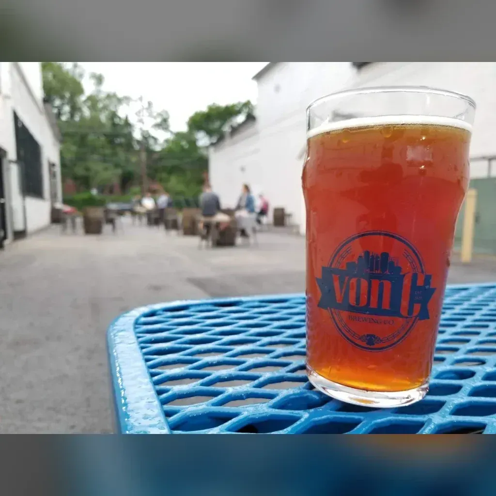 Branded von C Brewing pint of golden Pig Dog Pale Ale on a blue patio table in the von C Brewing beer garden, customers seated at tables in the soft-focus background