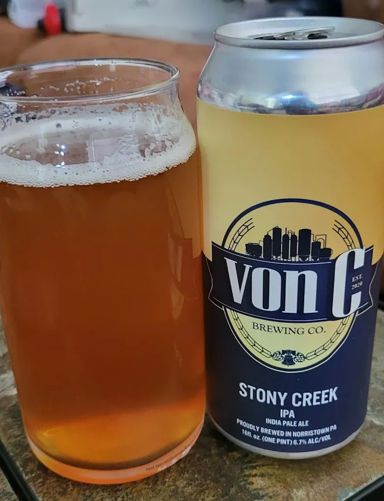 16 oz Stony Creek East Coast IPA can — von C Brewing wordmark, Norristown PA — beside a clear can-shaped glass with the freshly poured IPA on a stone bar top