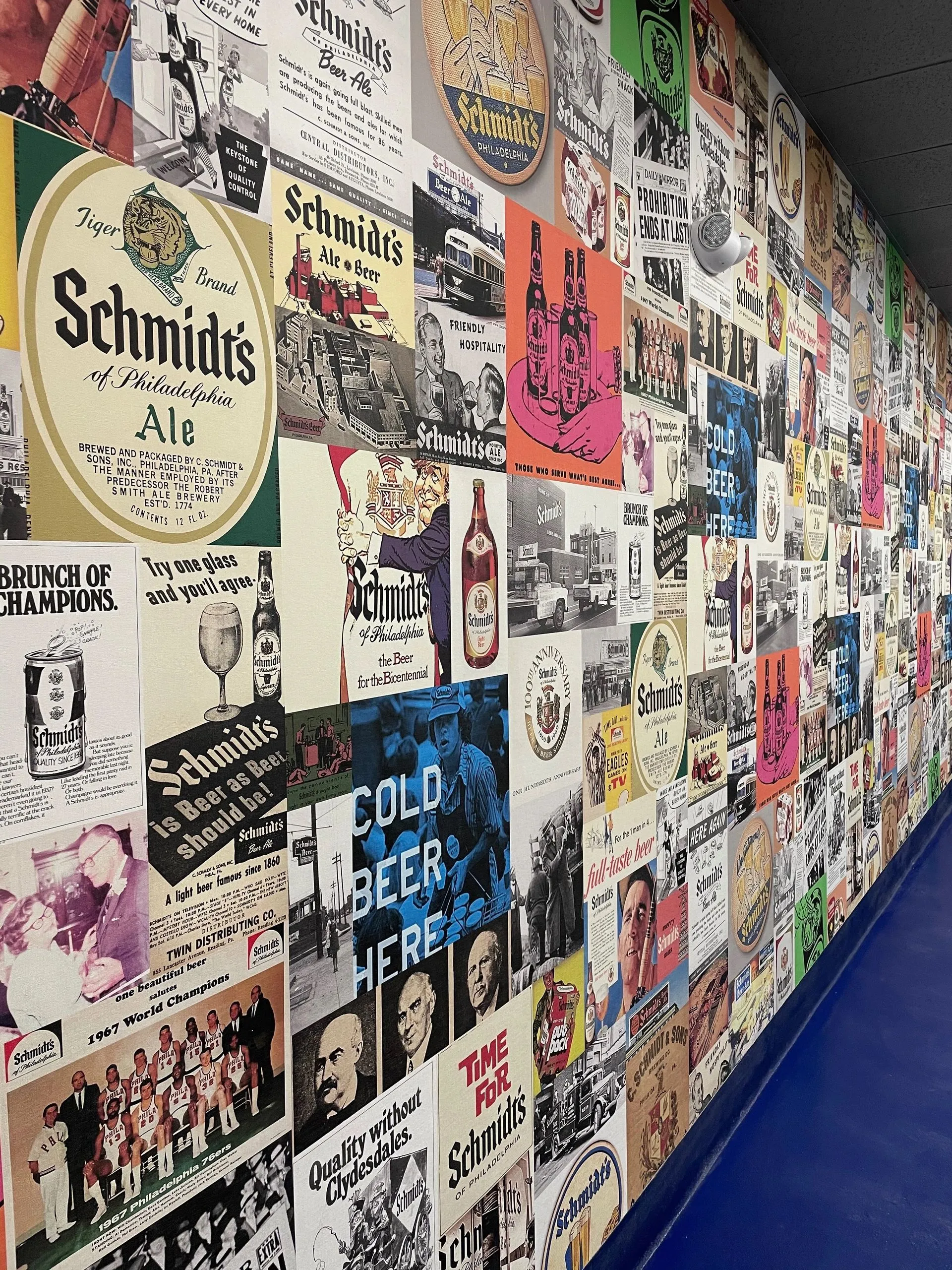 Wall of collaged vintage Schmidt's brewery advertisements inside the von C Brewing Co. taproom — BRUNCH of CHAMPIONS and TIME FOR Schmidt's archival posters arranged salon-style at bar height, the family record restored at scale in the room where their descendants pour again