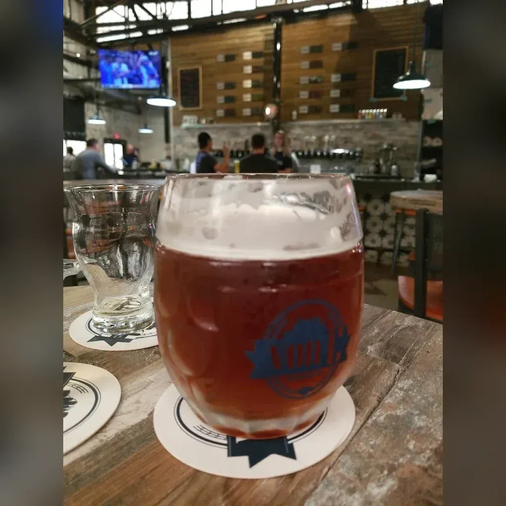 Copper-amber pint of That Much Further West Coast IPA in a von C Brewing branded mug with a thick cream head, taproom floor and bar visible in the soft background
