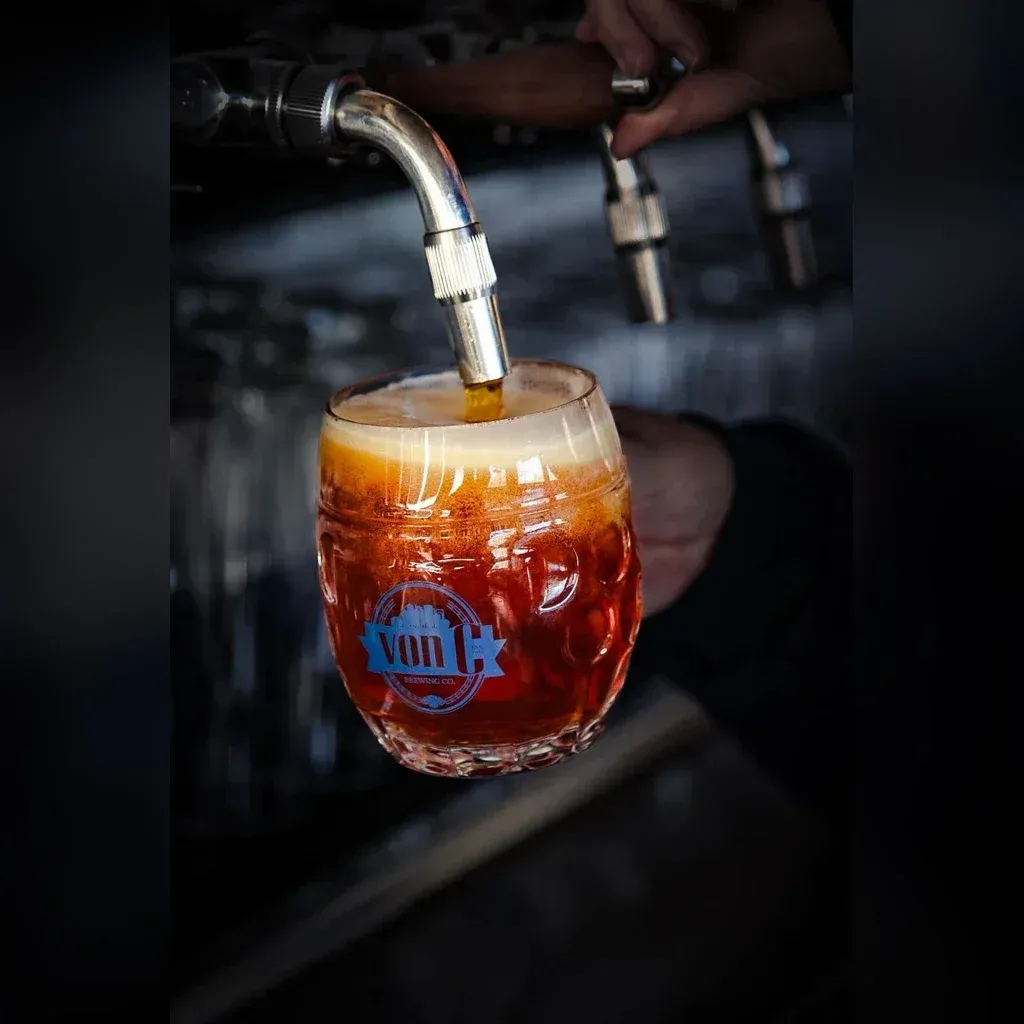 Side-pull faucet pouring a copper-colored Vienna lager into a custom von C Brewing dimpled mug, thick cream head forming, traditional Czech-style technique