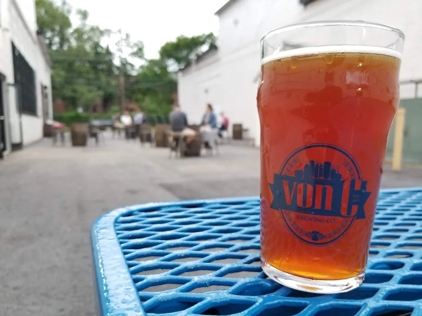 Vienna lager pint in a custom vonC pint glass on a blue mesh table in the von C Brewing Co. beer garden, with a group of guests seated at picnic tables in the background between the reclaimed-warehouse buildings on Stanbridge Street in Norristown