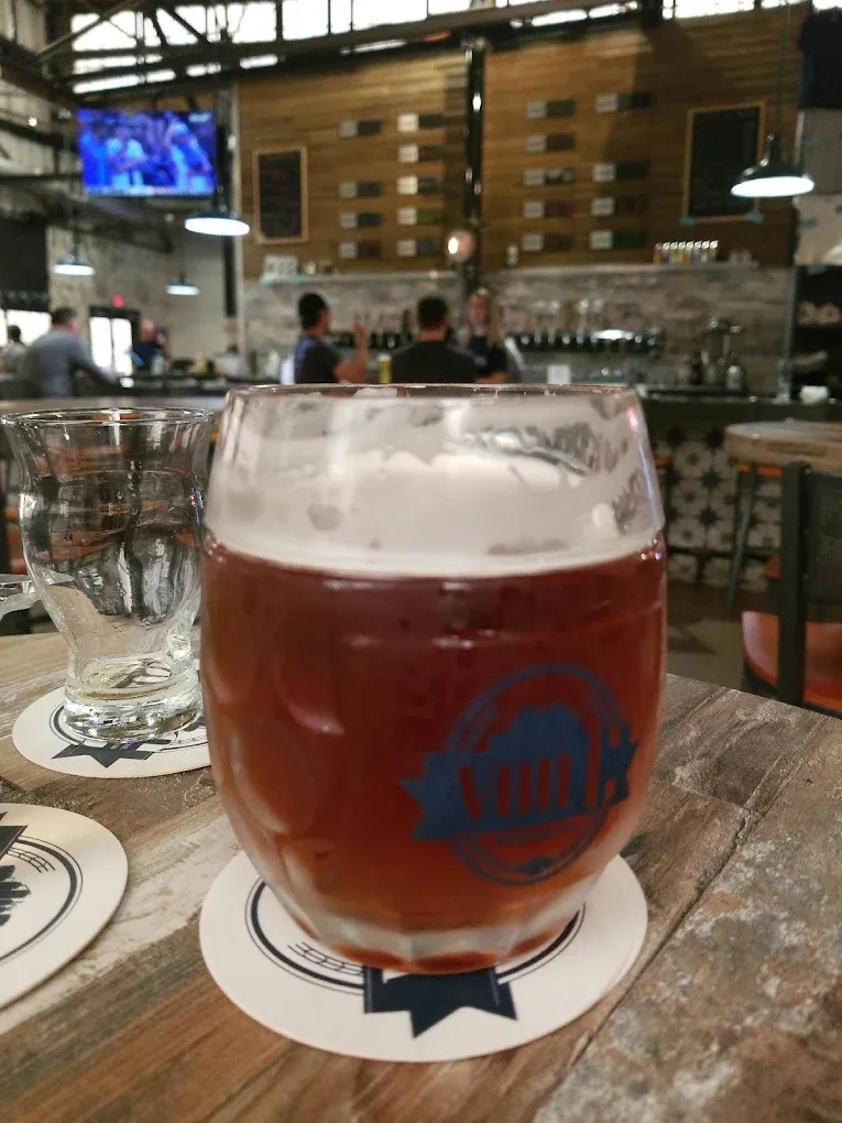 Copper-amber pint of That Much Further West Coast IPA in a von C Brewing branded mug with a thick cream head, taproom floor and bar visible in the soft background