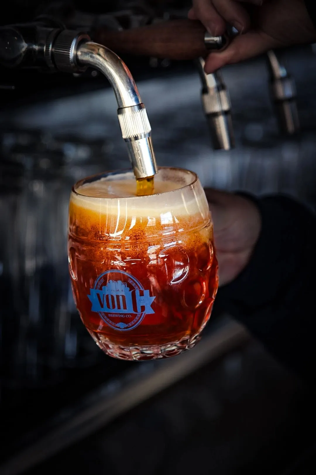 Side-pull faucet pouring copper-colored Vienna lager into a custom von C dimpled mug, creating a thick cream head — the traditional Czech-style technique used on every lager pour