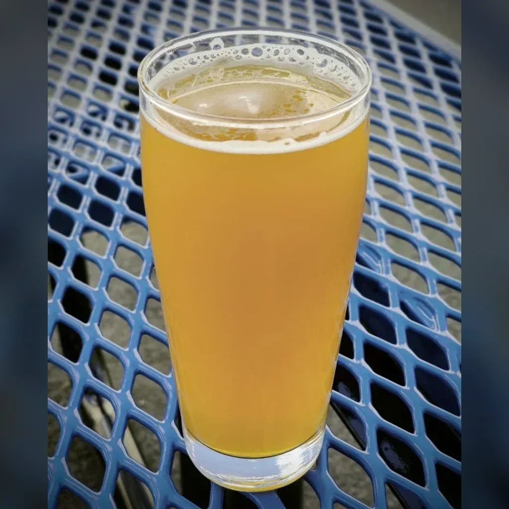 Tall pilsner glass of Walking on Clouds hazy yellow NEIPA on a bright blue perforated patio table at the von C Brewing beer garden in Norristown PA