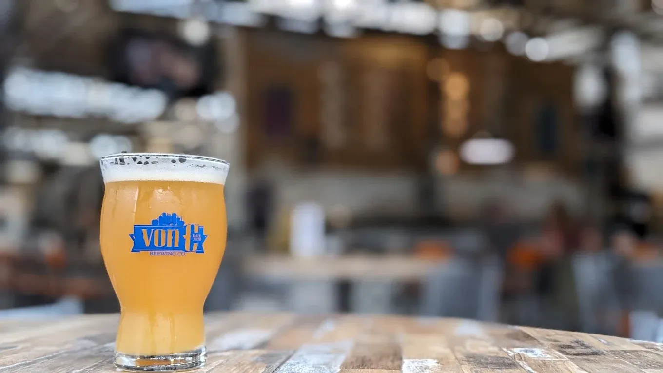 Close-up of a branded vonC Brewing pint glass of Walking on Clouds NEIPA on a reclaimed-wood bar top at the Norristown taproom — hazy yellow body, thin white head, the working brewhouse and Czech side-pull faucets soft-focused in the background
