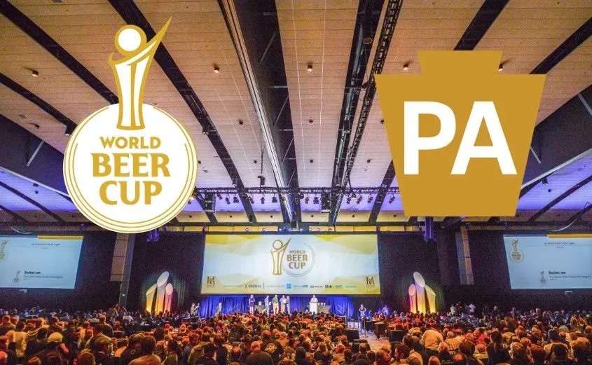 2023 World Beer Cup® awards ceremony — soaring convention-hall ceiling beams, theatrical stage lights, packed audience, the WBC trophy backdrop and Pennsylvania keystone overlaid as event branding